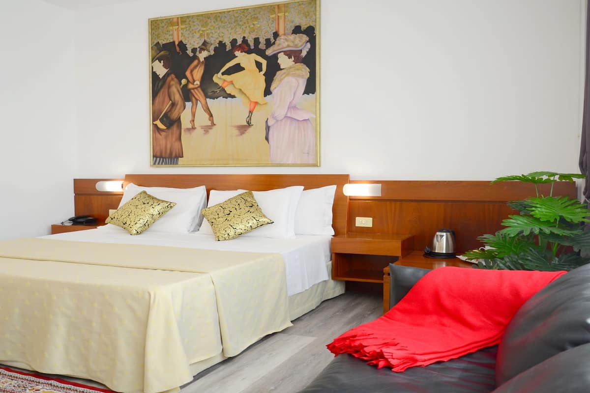 A hotel room with a large bed, a wooden headboard, two bedside tables, a red throw blanket, a black leather sofa, and a painting on the wall.