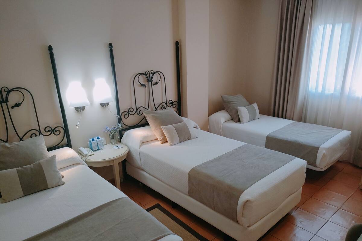 A hotel room with two single beds, a round bedside table with a lamp, a phone, and a vase with flowers.
