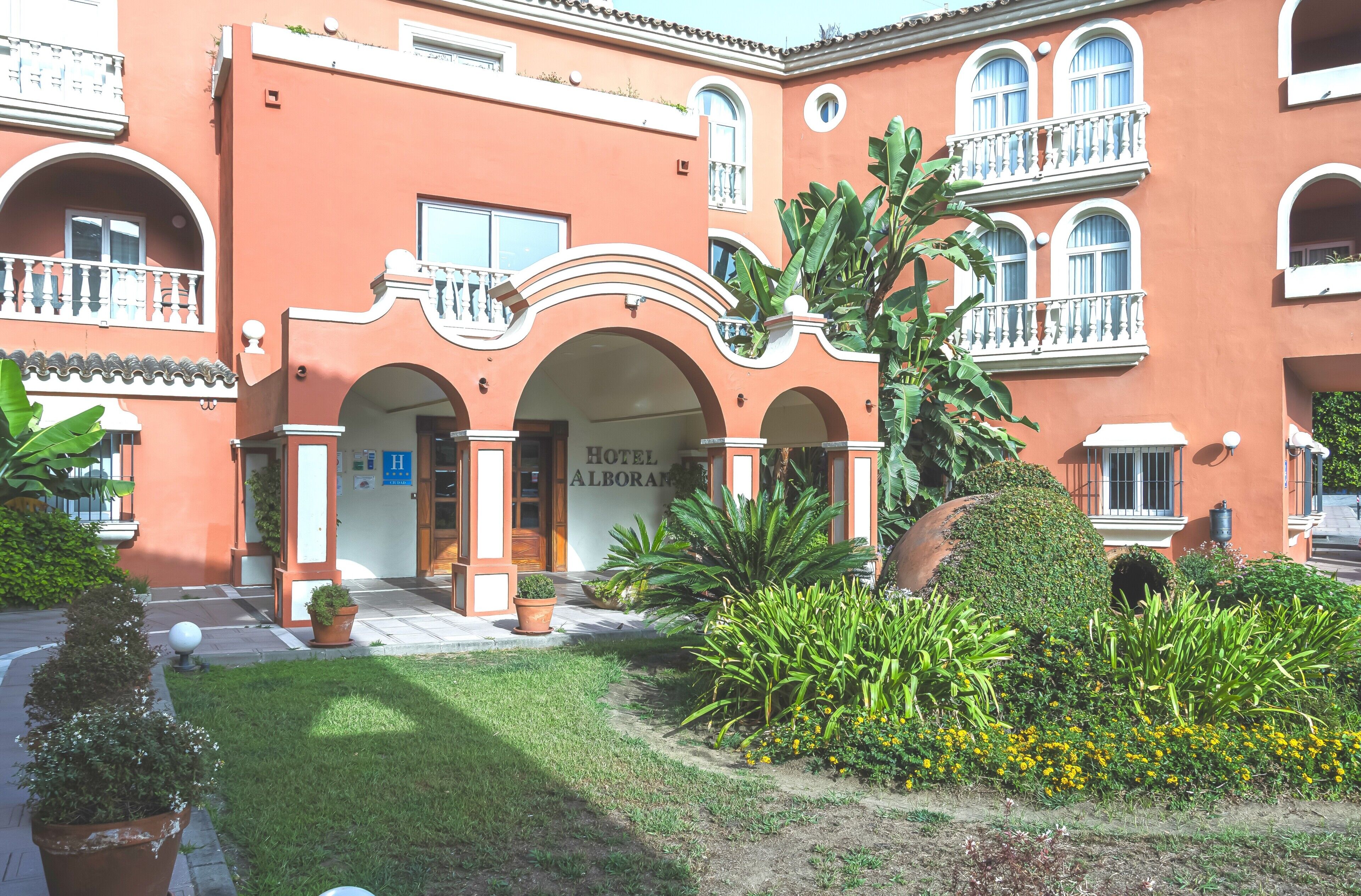 A hotel with a red facade, multiple balconies, and a well-maintained garden with various plants and flowers.