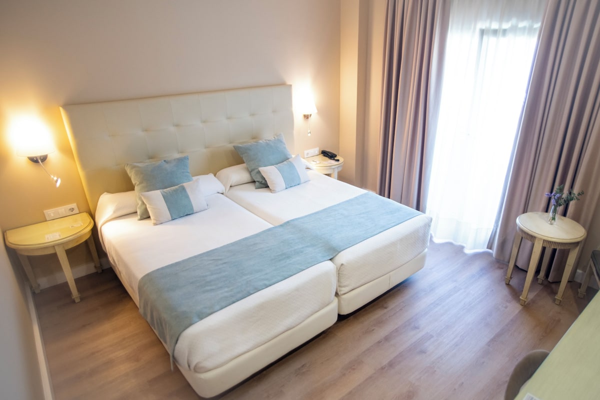 A double bed with a tufted headboard, two bedside tables with lamps, and a view of a window with curtains.