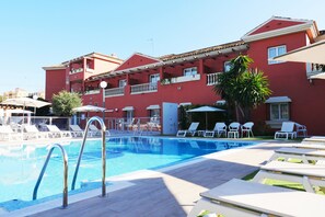 Piscina con sillas de descanso y sombrillas, rodeada por un edificio rojo con balcones.