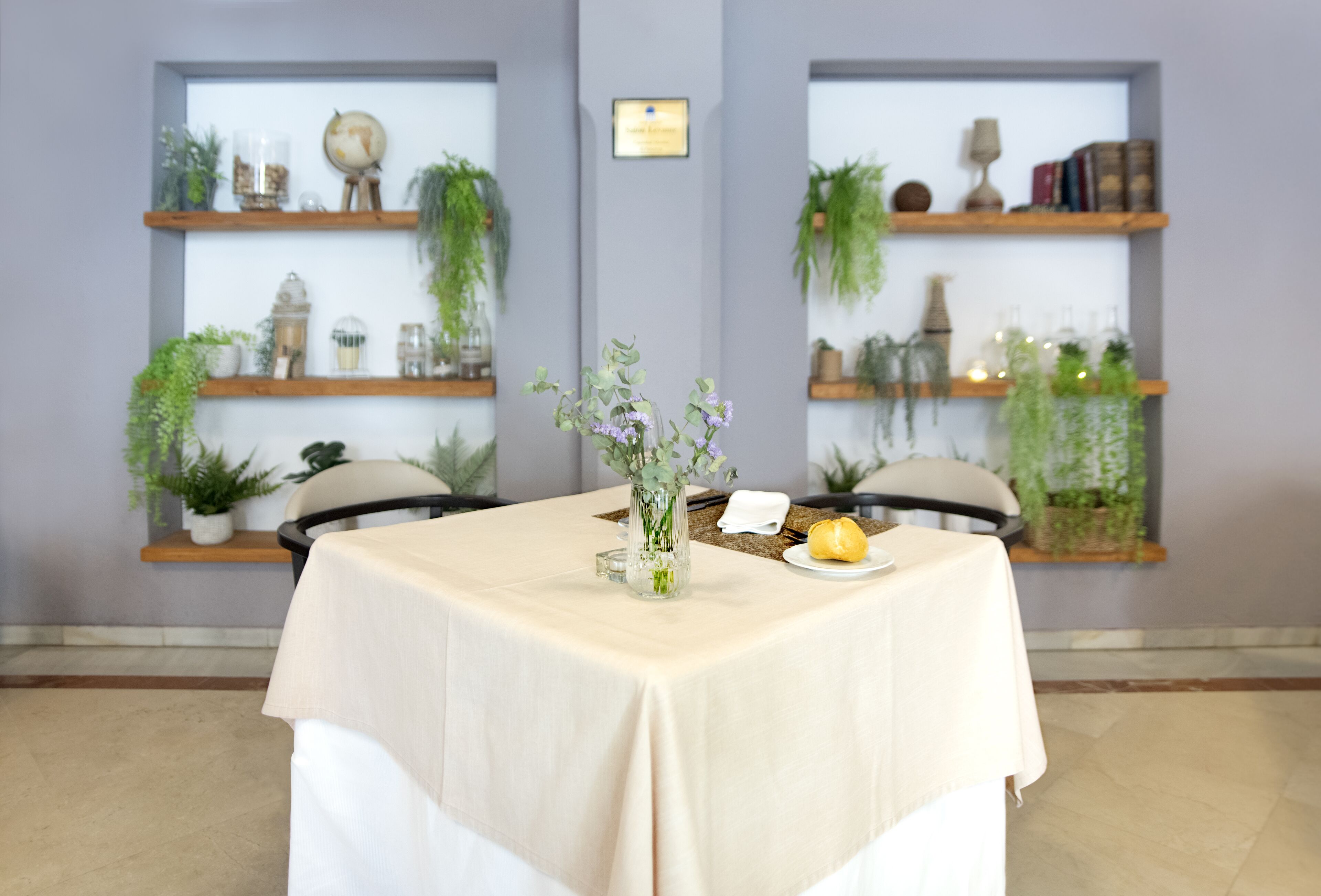 Un comedor con una mesa dispuesta para dos personas, rodeado de estantes con plantas y objetos decorativos.