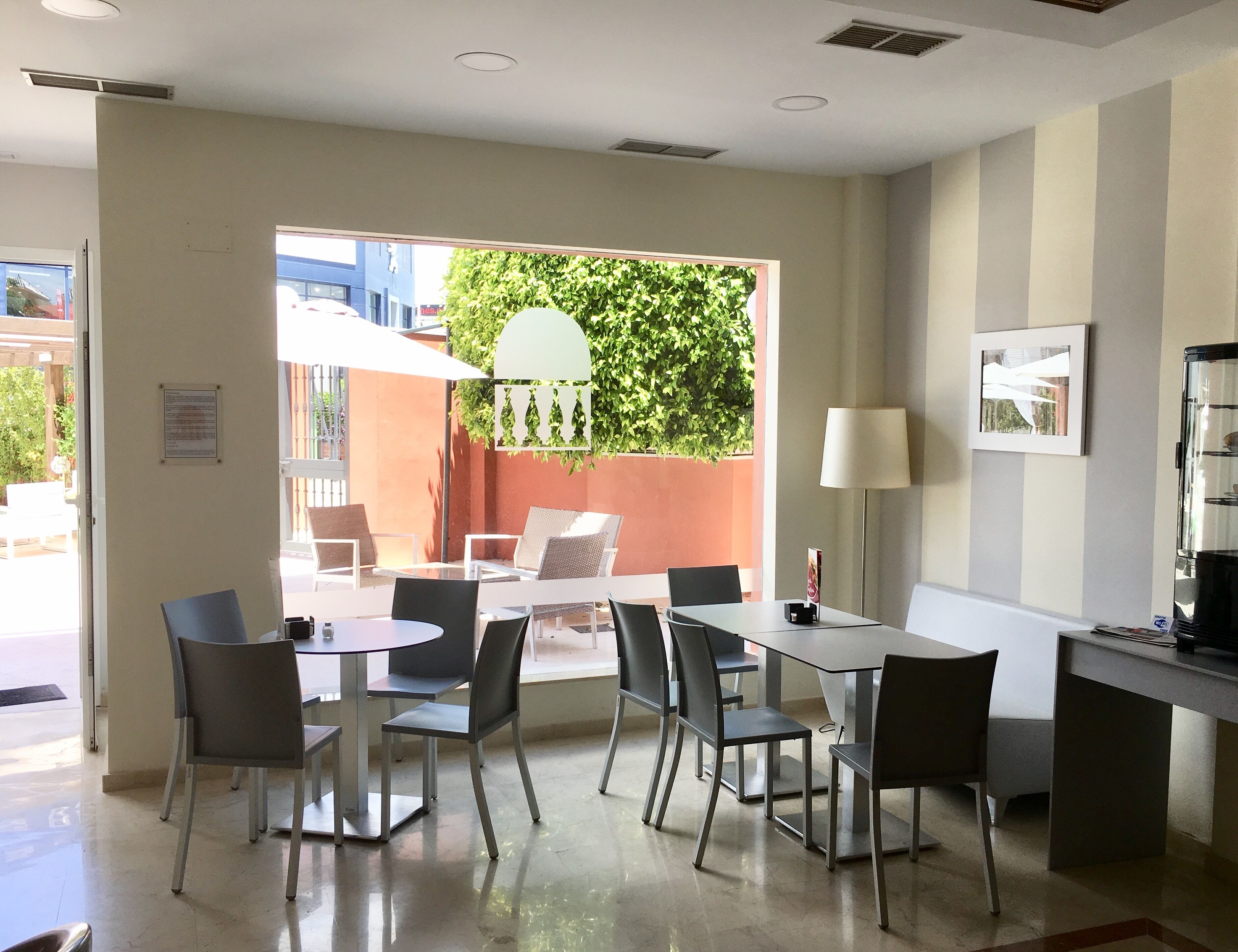 Una habitación de hotel moderna con zona de comedor que incluye una mesa, sillas, una lámpara y vistas a un área verde exterior a través de un ventanal.