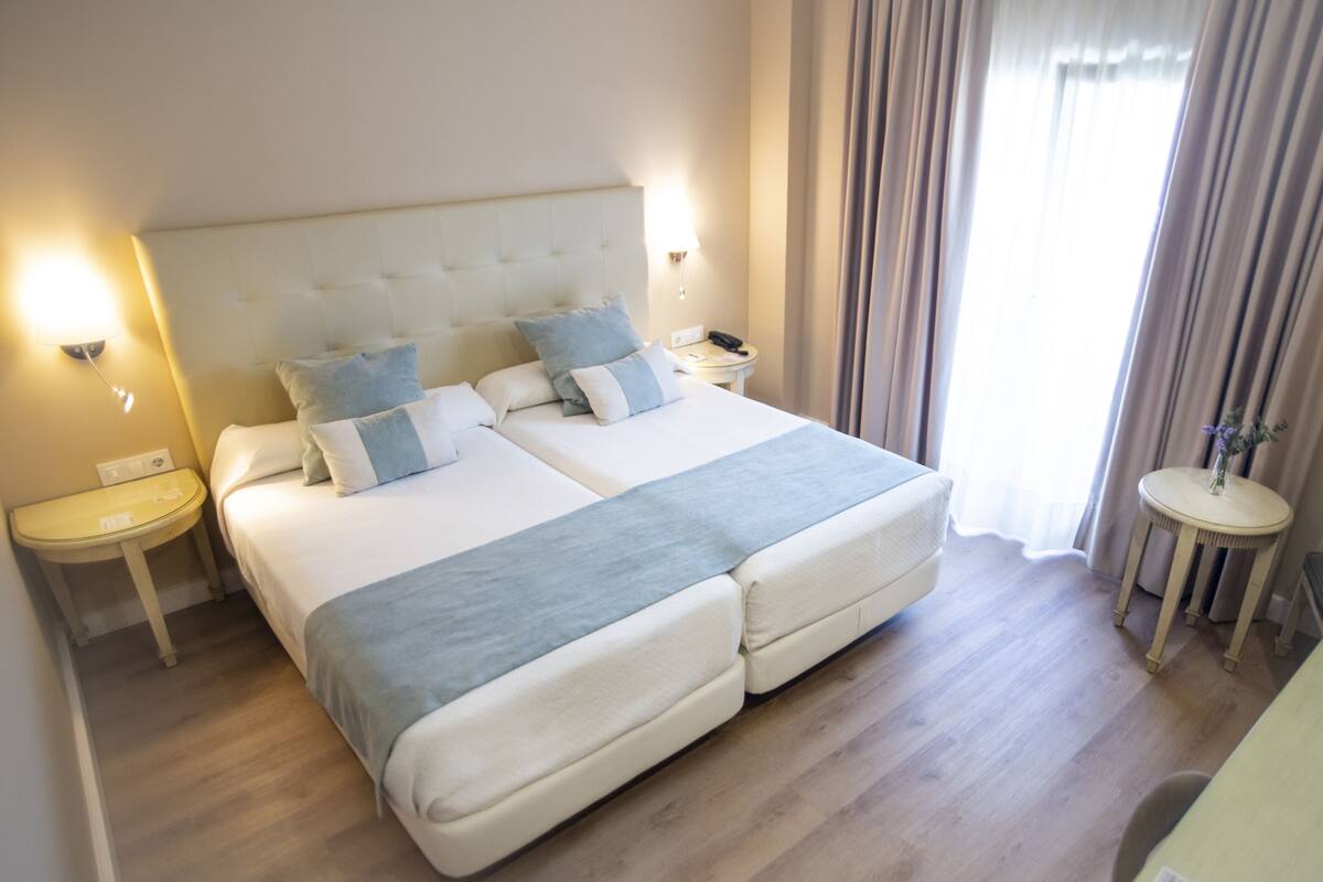 A double bed with a tufted headboard, two bedside tables with lamps, and a view of a window with curtains.