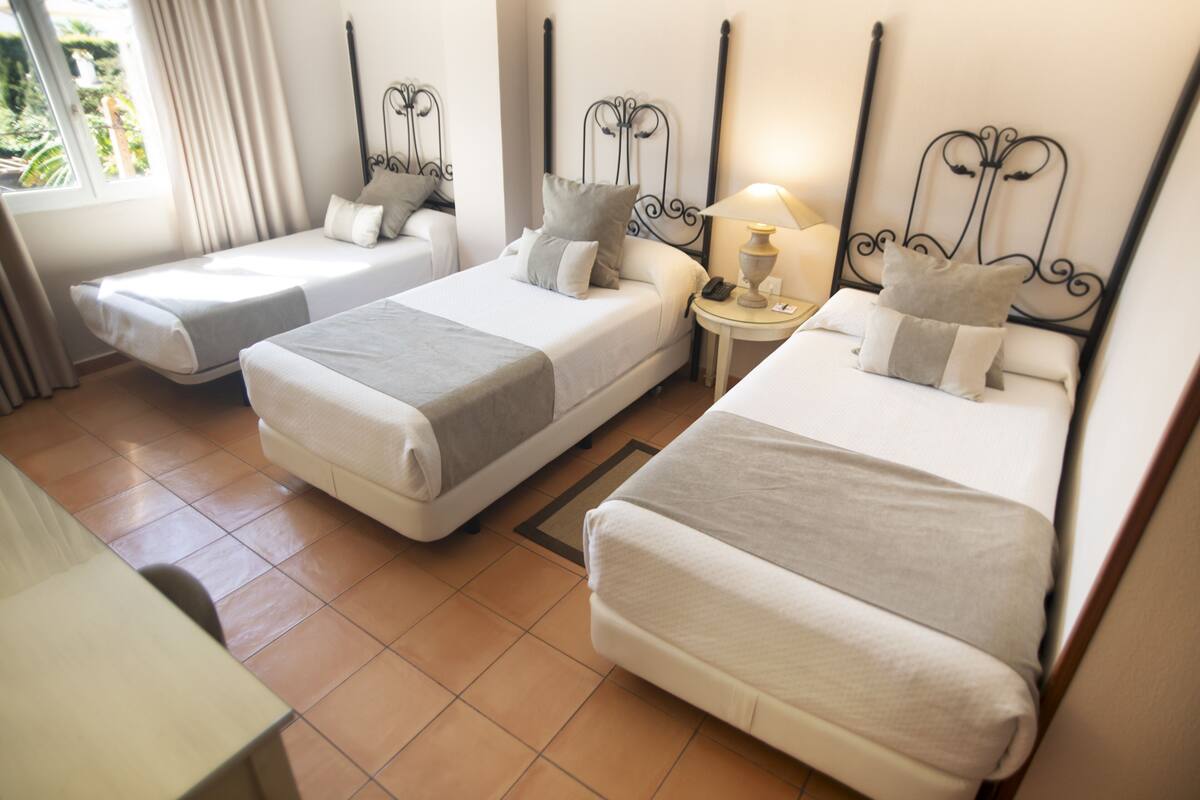 A hotel room with three single beds, a bedside table with a lamp, and a view of the outdoors.