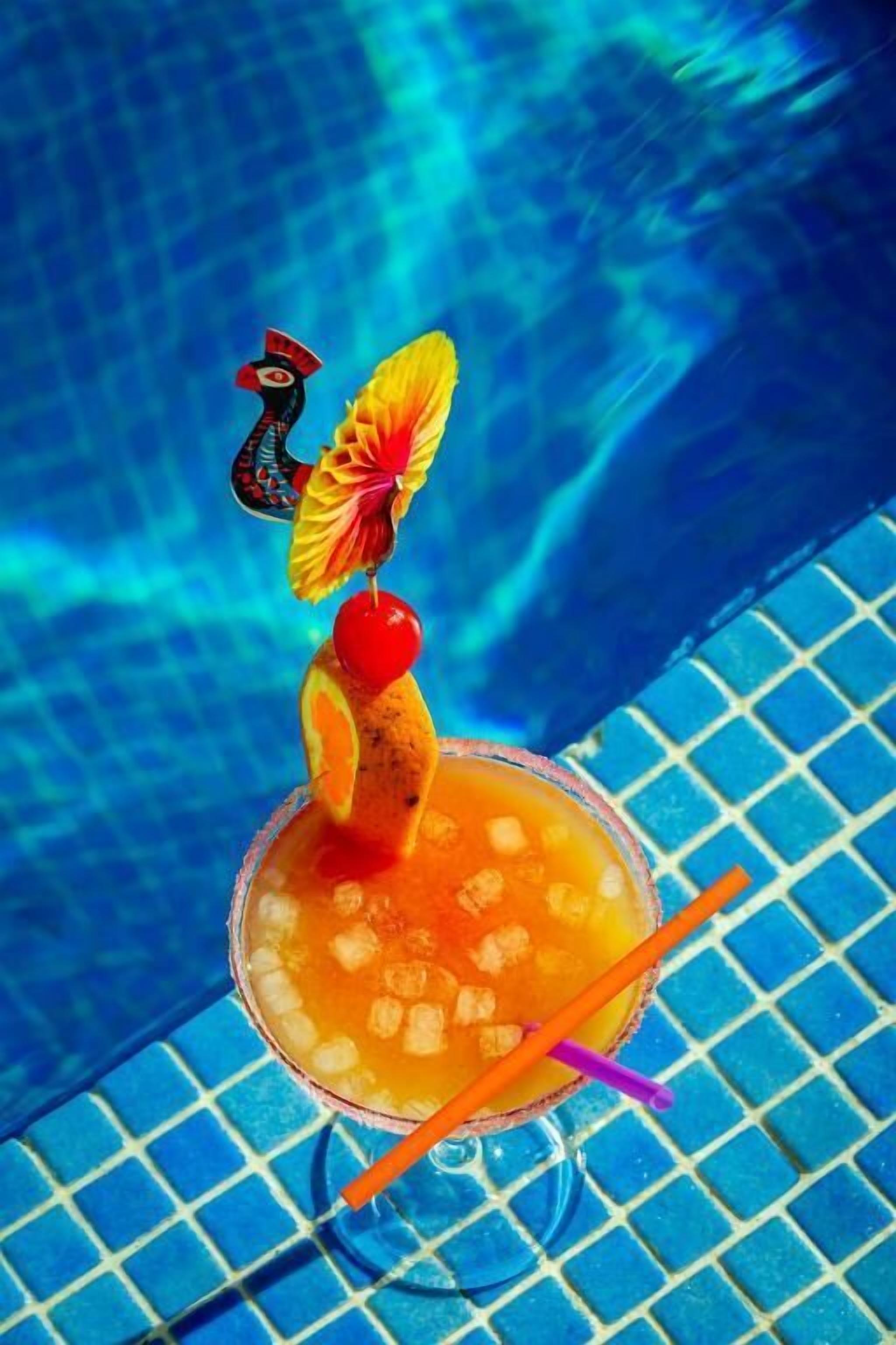 Un cocktail décoré d’un petit parapluie et d’une figurine, servi dans un verre rempli de glaçons, sur une surface bleue carrelée.