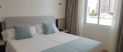 A hotel room with a bed, a nightstand, a lamp, a window with curtains, and a view of buildings outside.