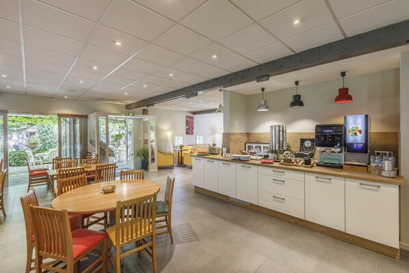A spacious dining area with wooden tables and chairs, a central island with a coffee machine, and a view of the outdoor garden.