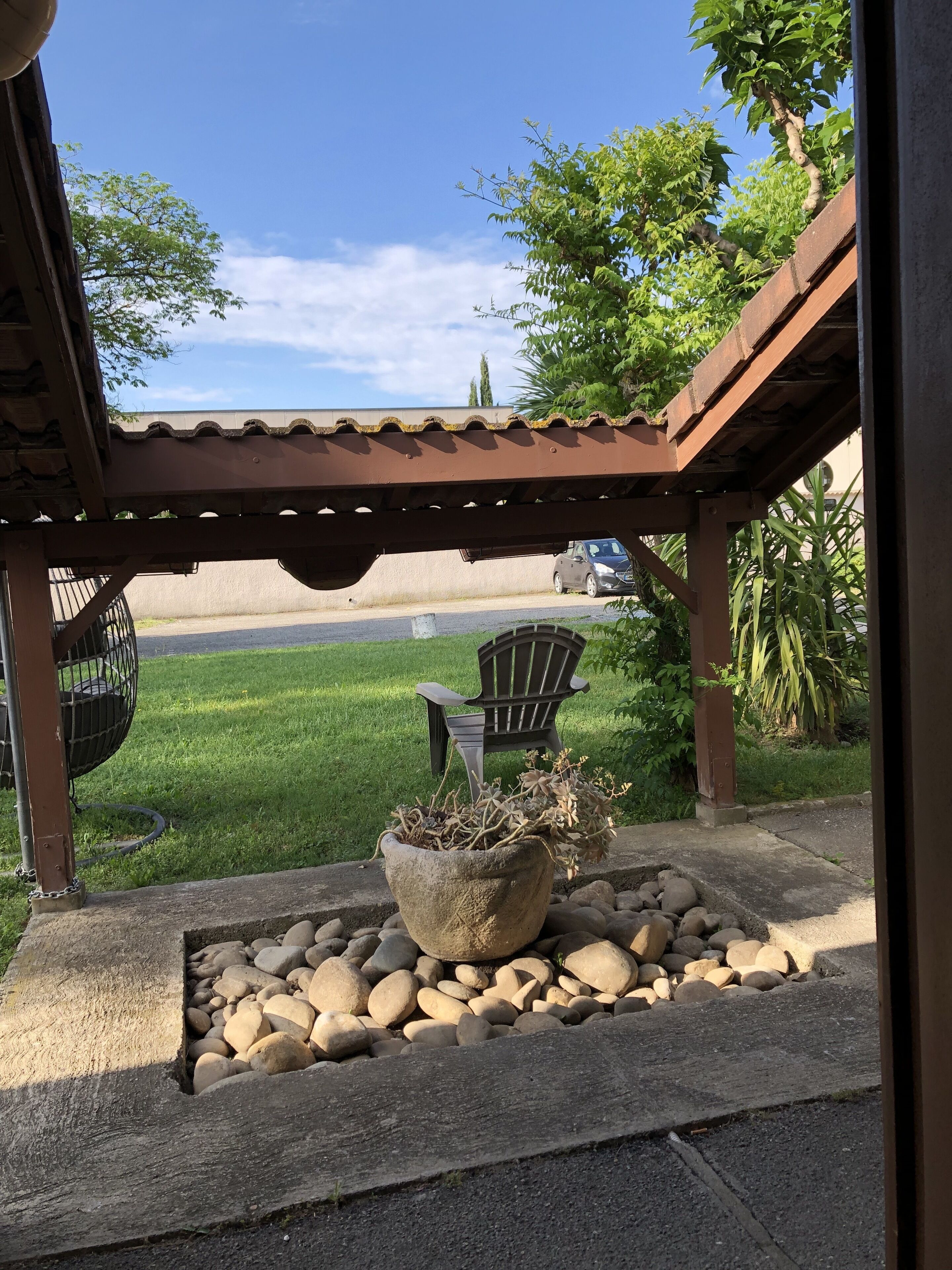 Ein überdachter Patio-Bereich mit Betonboden, einem Steinpflanzgefäß mit Pflanze, einem schwarzen Stuhl und einem Metallstuhl.