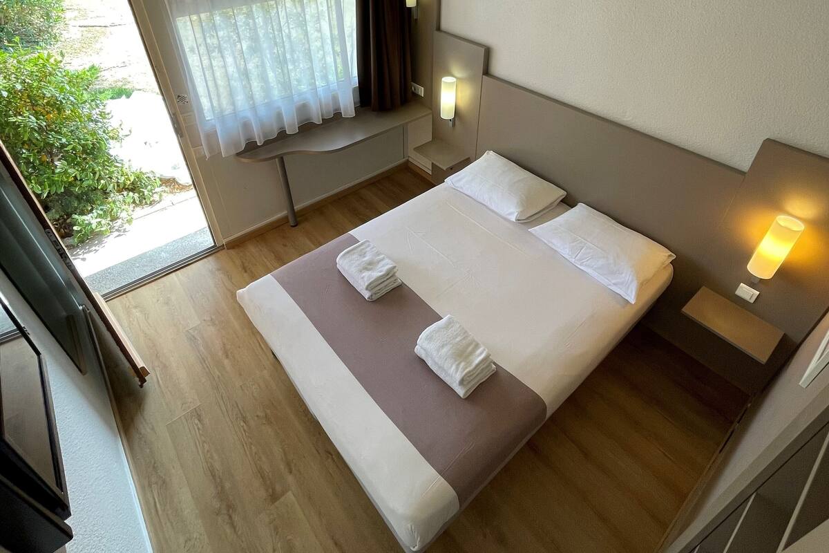 A hotel room with a large bed, two bedside tables, a window with curtains, and a view of greenery outside.