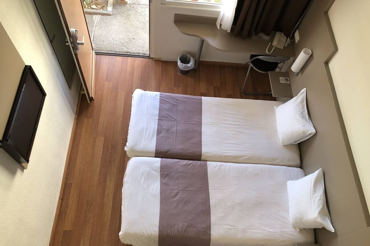 A hotel room with two beds, a wooden floor, a window, a trash bin, and a phone on the wall.