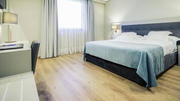 A hotel room with a bed, a desk, a chair, a lamp, and a window with curtains.