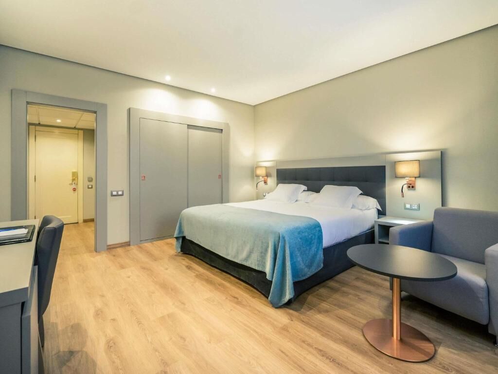 A modern hotel room with a bed, a desk, a chair, and a small table.