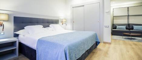 A modern hotel room with a large bed, bedside tables, a wardrobe, and a seating area with a sofa and chairs.