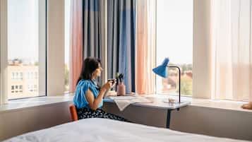 A hotel room with a bed, a desk with a lamp, a window with curtains, and a view of buildings outside.