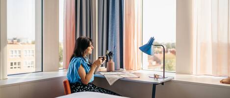 A hotel room with a bed, a desk with a lamp, a window with curtains, and a view of buildings outside.