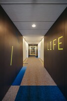 Un couloir doté d’un sol recouvert de moquette, d’un tapis bleu courant le long du couloir, et de murs sur lesquels le mot « VIE » est illuminé en jaune.