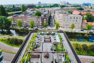 Un jardin sur le toit, agrémenté de bancs et de tables en bois, entouré d’immeubles et bordé par une rivière.