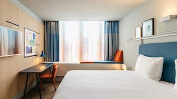 Habitación de hotel con cama, escritorio, silla, ventana con cortinas y estante en la pared con fotografías.