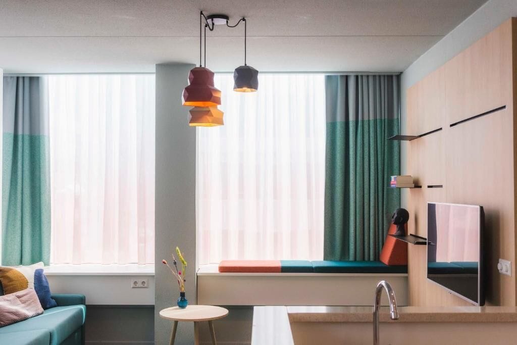 A modern hotel room with a sofa, a small round table, a window seat with curtains, a TV mounted on the wall, and a sink.