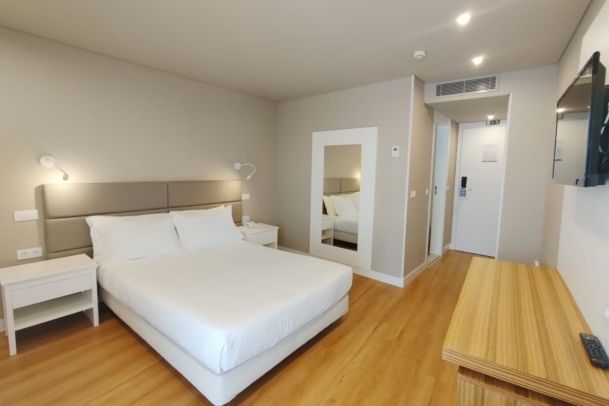 A modern hotel room with a large bed, bedside tables, a mirror, a TV, and a wooden floor.