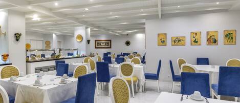 A restaurant with white tables and blue chairs, a kitchen in the background, and artwork on the walls.