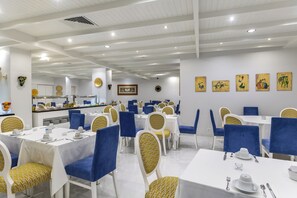 A restaurant with white tables and blue chairs, a kitchen in the background, and artwork on the walls. - Hotel Cristal Caldas (Caldas da Rainha)