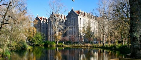 Un edificio histórico con un estanque grande y reflectante en el frente, rodeado de árboles y una terraza de madera.