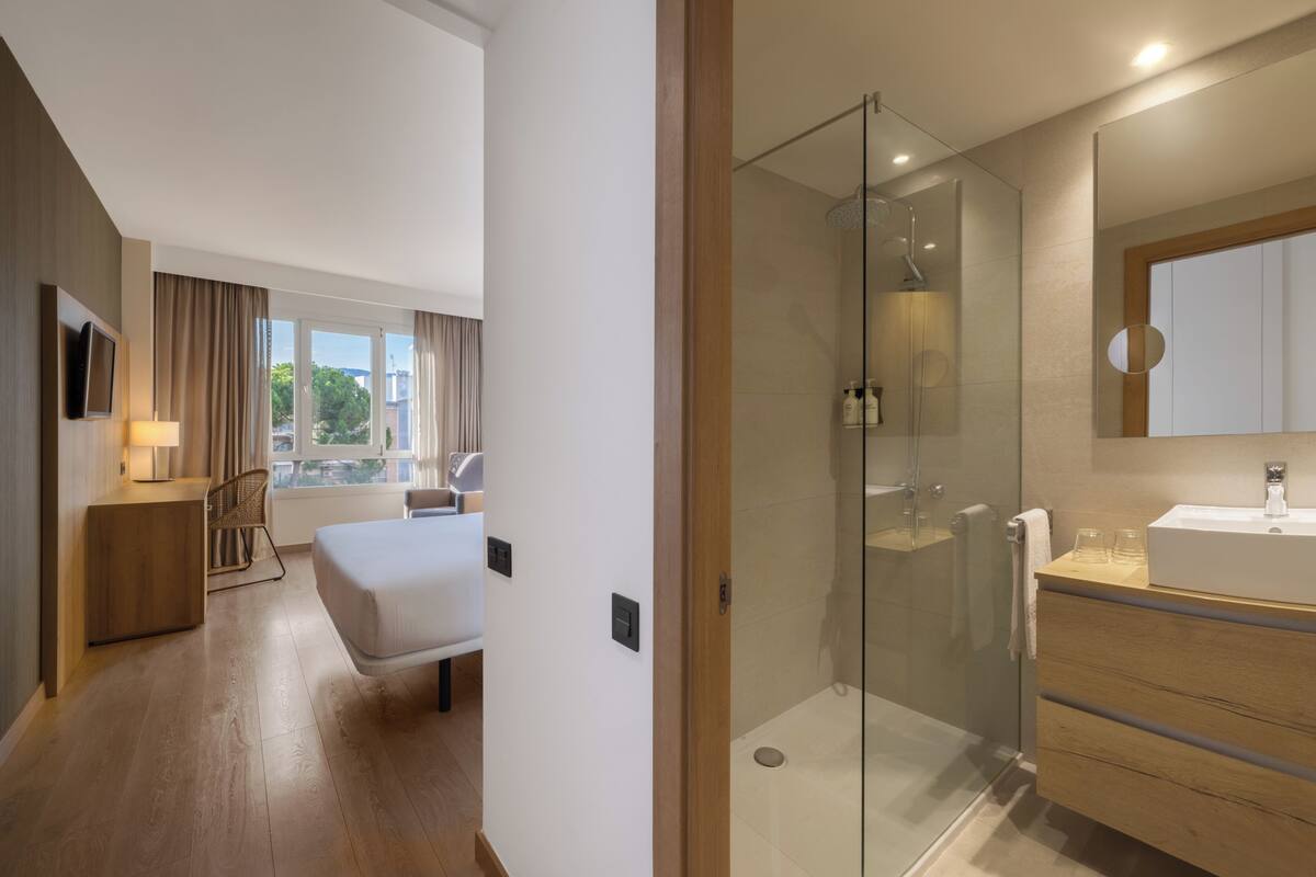 Habitación de hotel moderna con una cama, una mesita de noche, un televisor, ducha con mampara de vidrio y vistas al exterior.