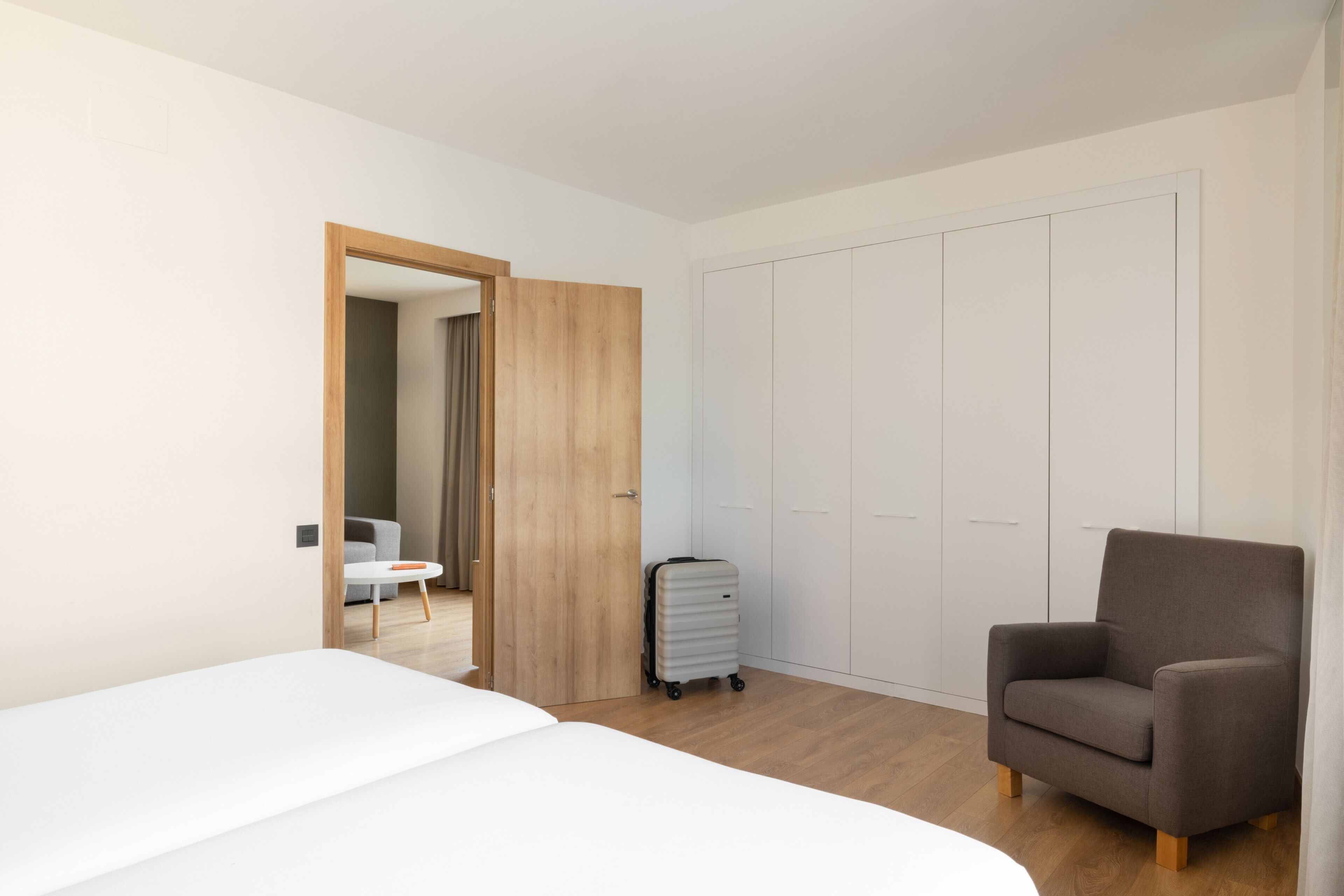 A hotel room with a bed, a chair, a suitcase, a wardrobe, and a door leading to another room.
