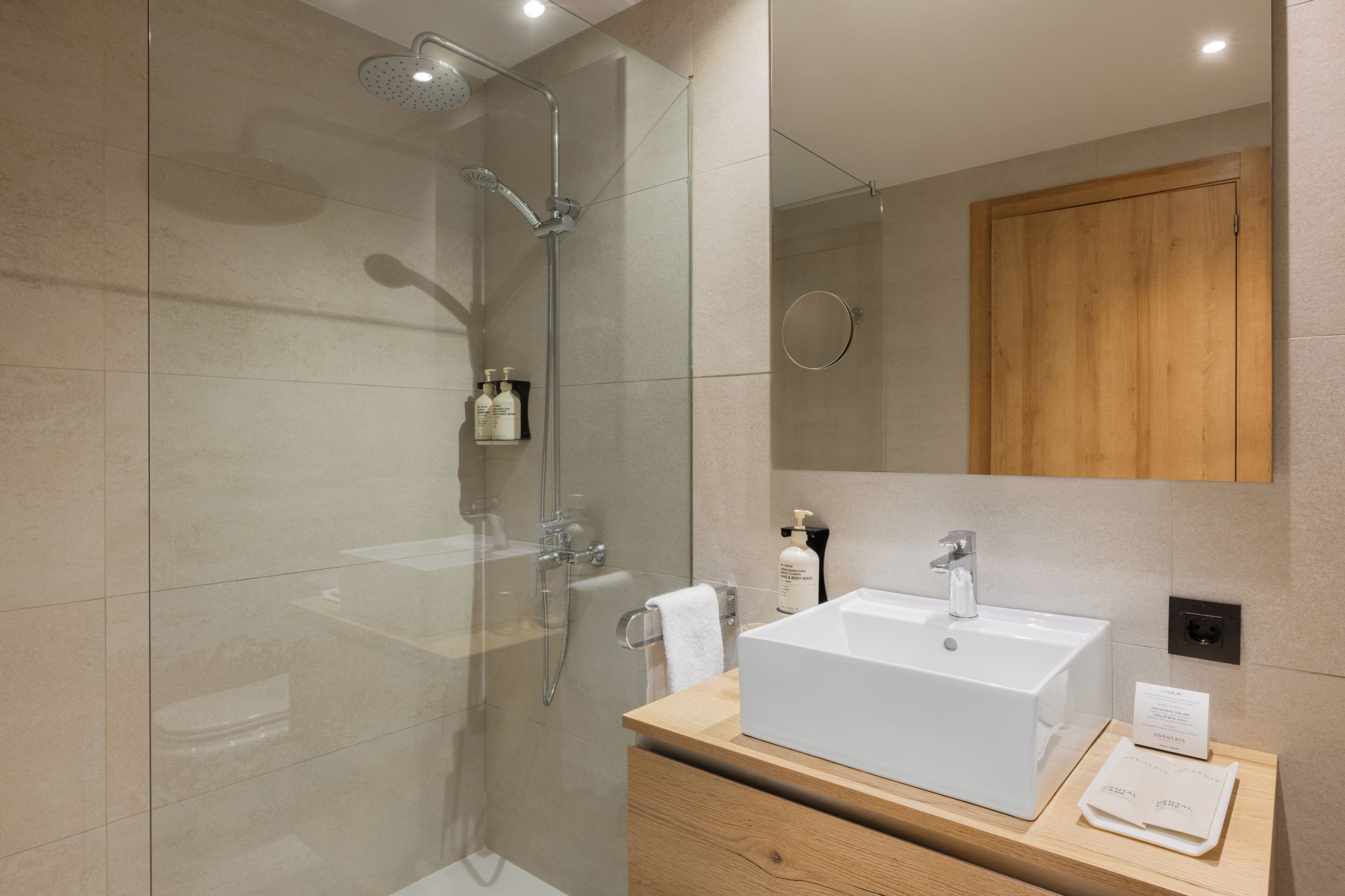 Un baño moderno con ducha de vidrio, tocador de madera con lavabo blanco y un espejo sobre el lavabo.