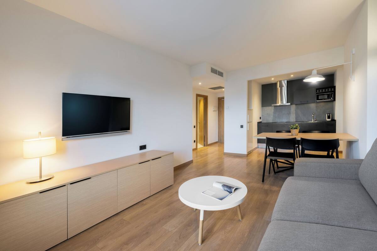 Una sala moderna con un televisor de pantalla plana, una mesa de centro redonda, un sofá y un comedor con una cocina al fondo.