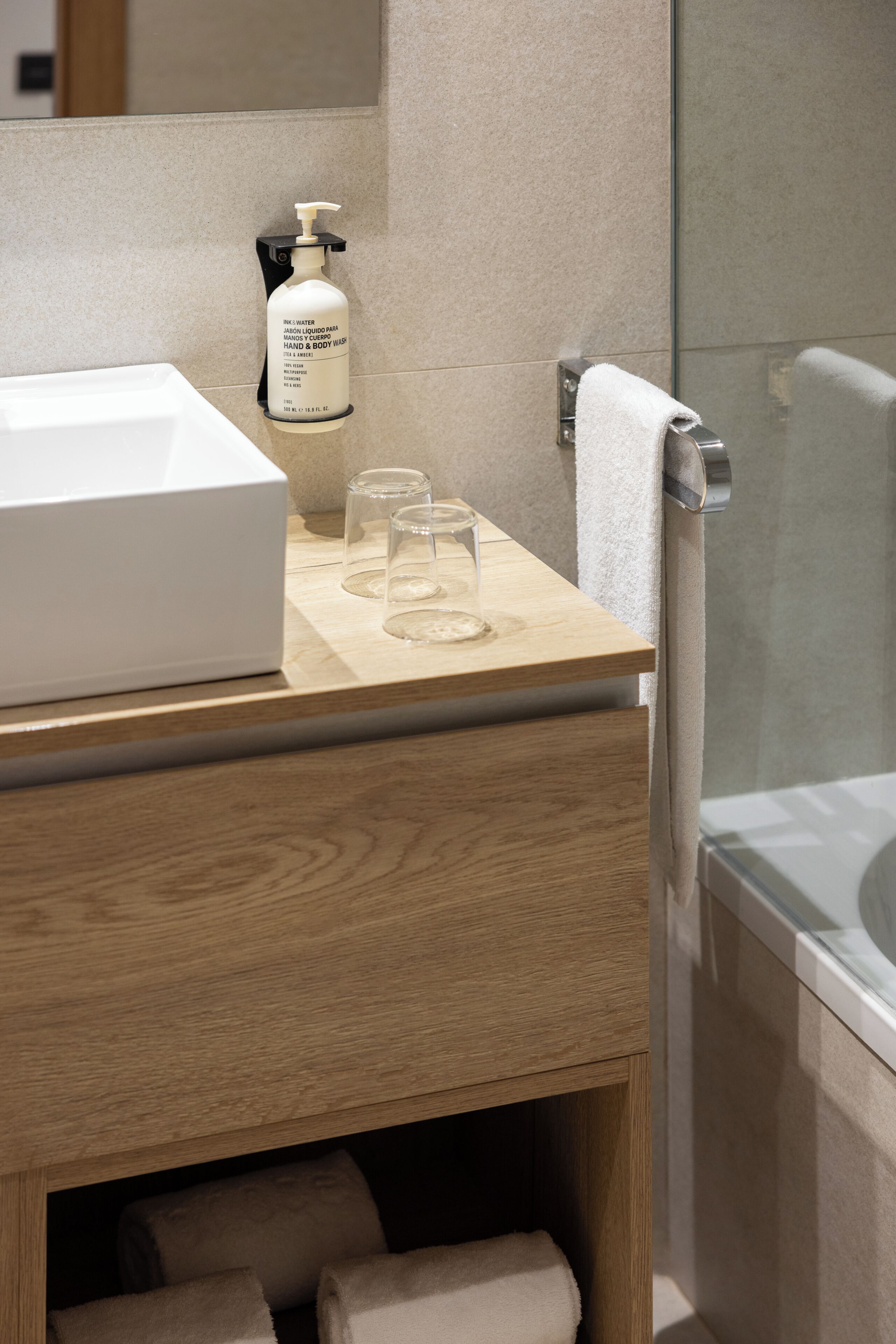 A bathroom with a white sink, a bottle of hand and body wash, two glasses, a towel, and a glass shower enclosure.