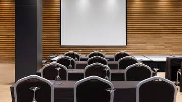 Un salón de conferencias con una pantalla grande, varias mesas con sillas y un proyector.