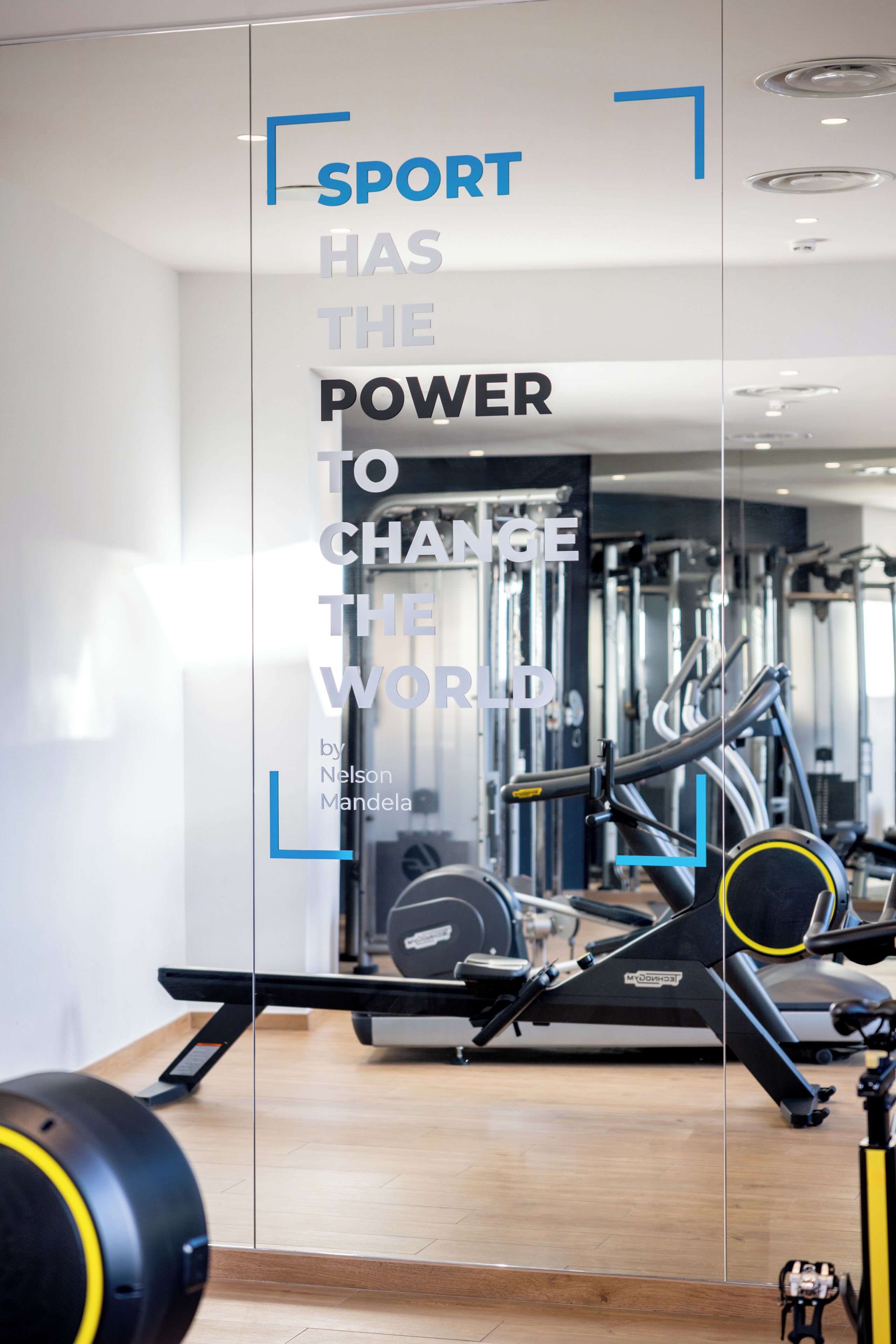 Un gimnasio con equipo de ejercicio, una frase motivacional en la pared y el reflejo del equipo en la pared de vidrio.