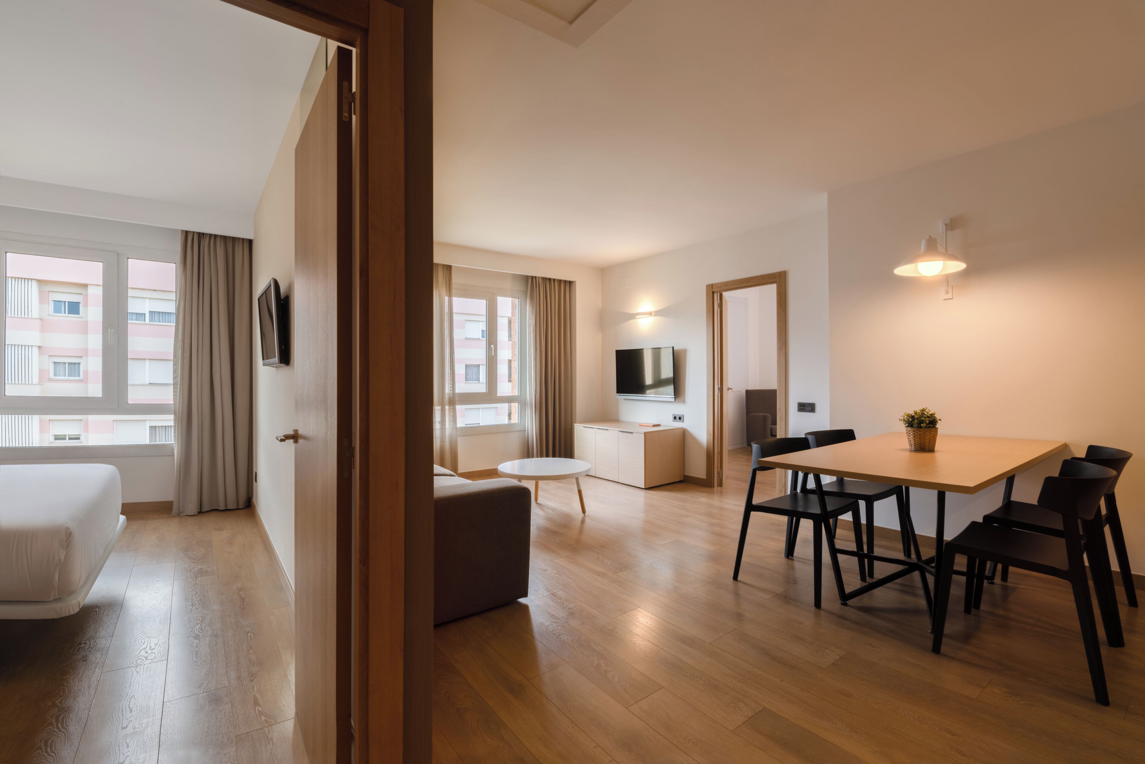 A modern hotel room with a wooden floor, a bed, a dining area with a table and chairs, a TV, and a kitchen area.