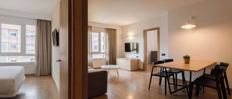 A modern hotel room with a wooden floor, a bed, a dining area with a table and chairs, a TV, and a kitchen area.