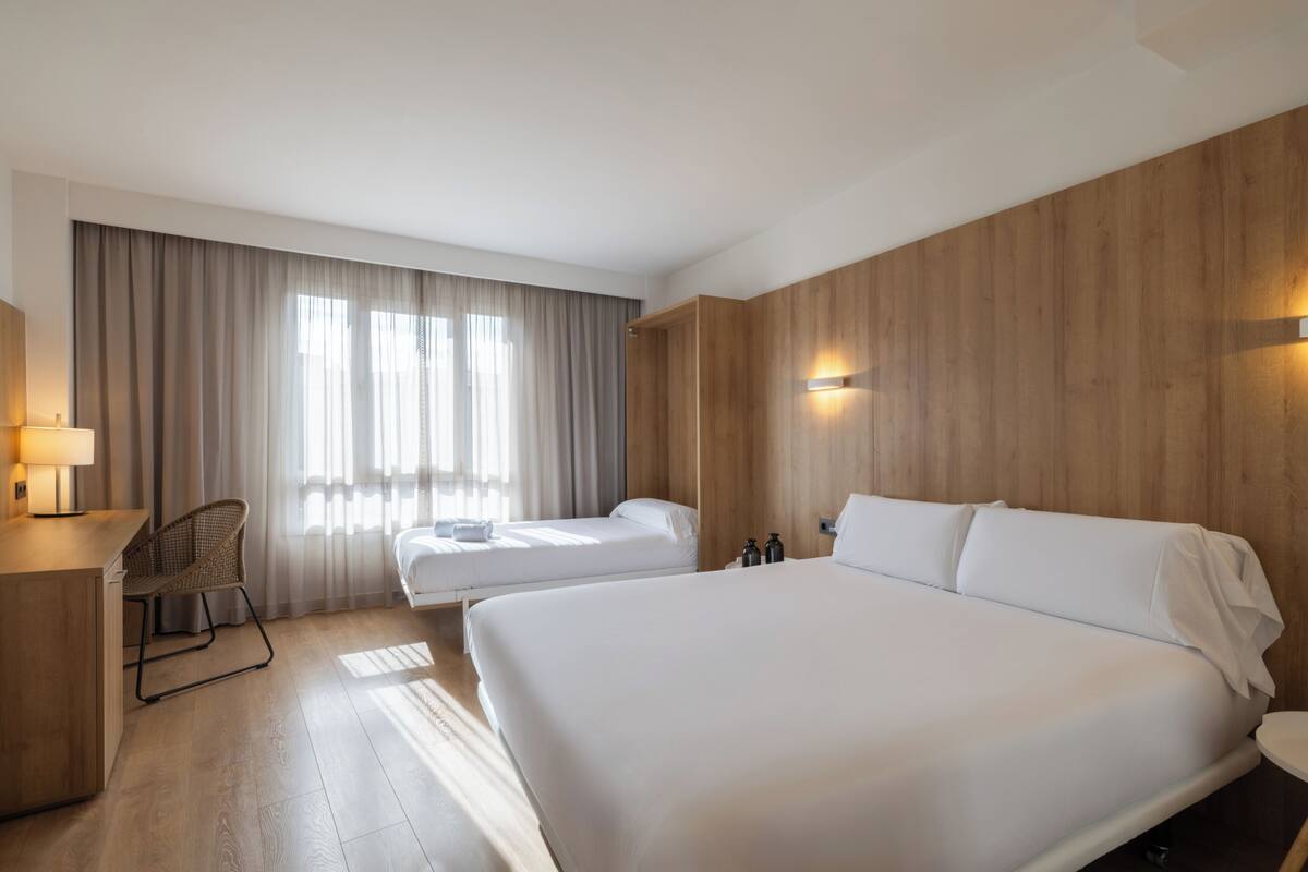 Una habitación de hotel con una cama grande, una cama más pequeña, un escritorio, una silla y un ventanal con cortinas.