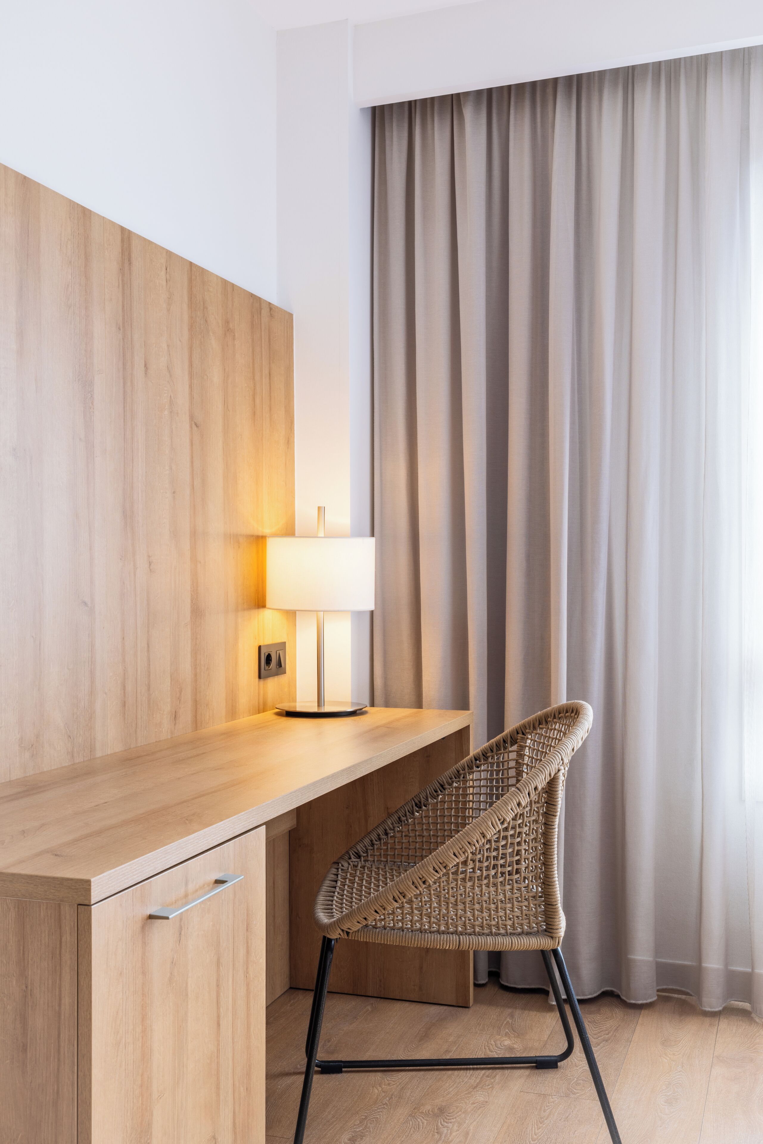 Una habitación moderna con un escritorio de madera, una silla de mimbre, una lámpara y un amplio ventanal con cortinas.