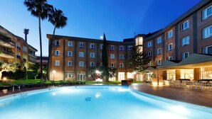 Ein Hotelpoolbereich mit blauem Himmel, Palmen und einem Gebäude mit mehreren Fenstern und Balkonen.