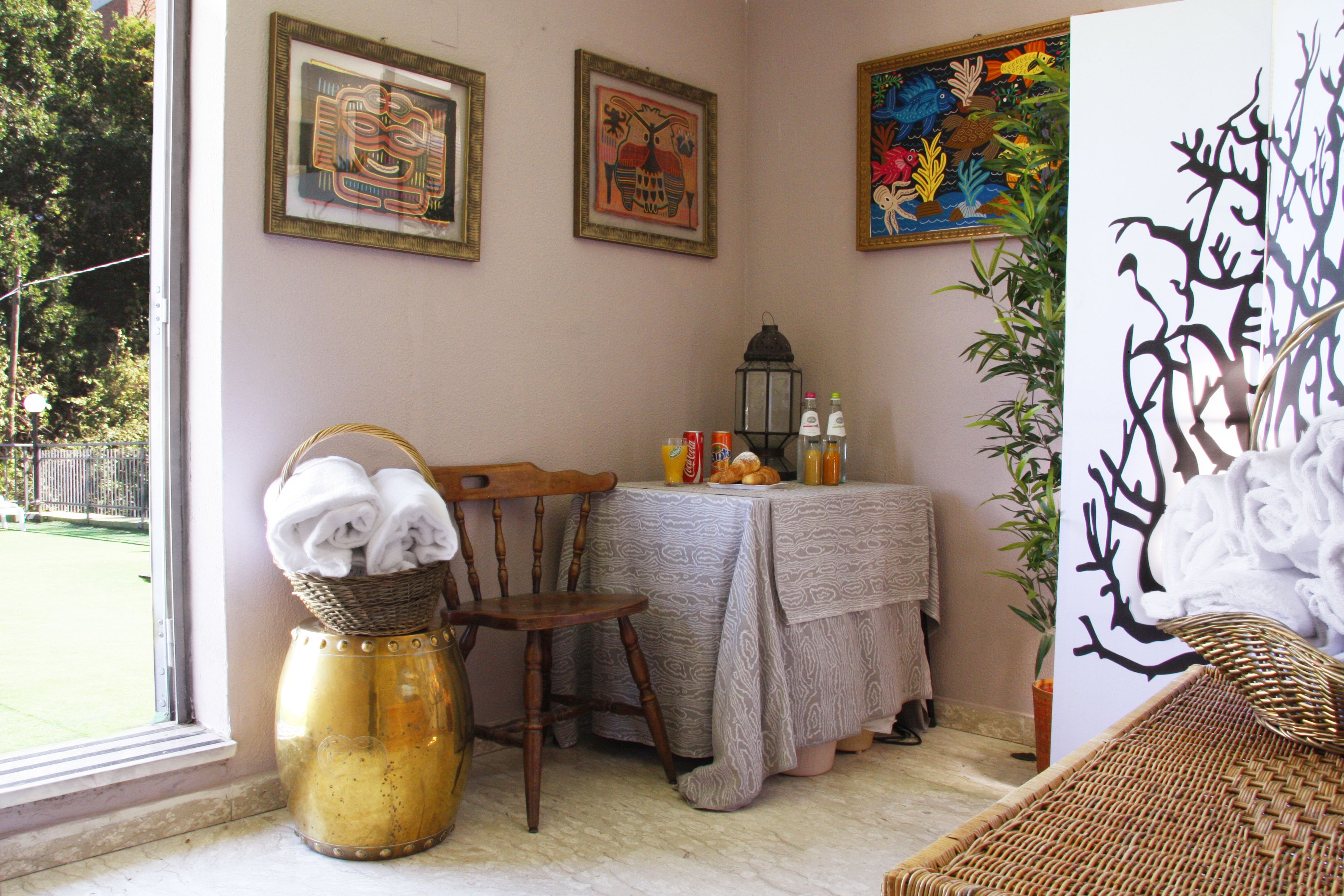 A room with a wicker chair, a table with a tablecloth, a vase with towels, and framed artwork on the wall.