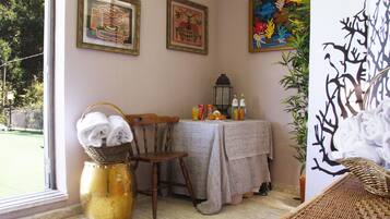 A room with a wicker chair, a table with a tablecloth, a vase with towels, and framed artwork on the wall.