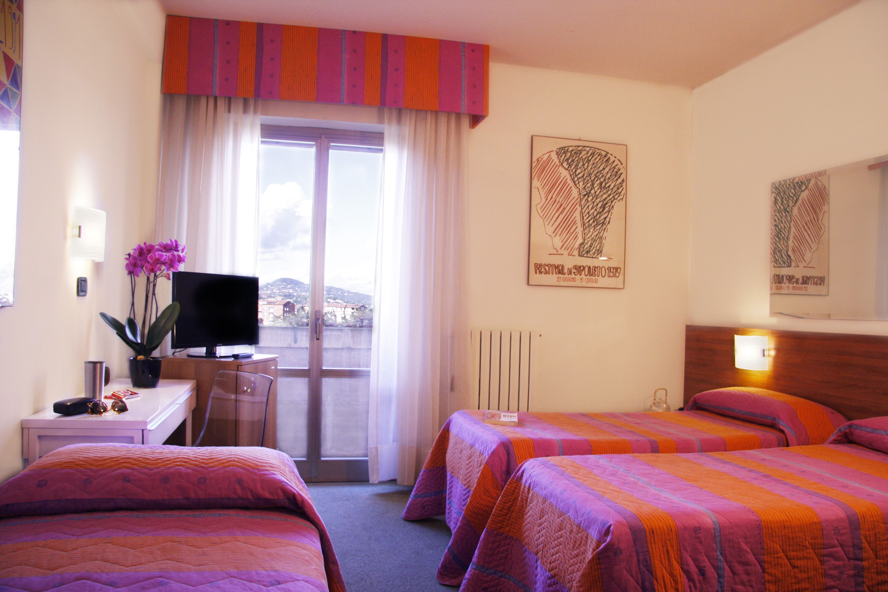 A hotel room with two beds, a desk with a TV, a potted plant, and a view of the city through a large window.
