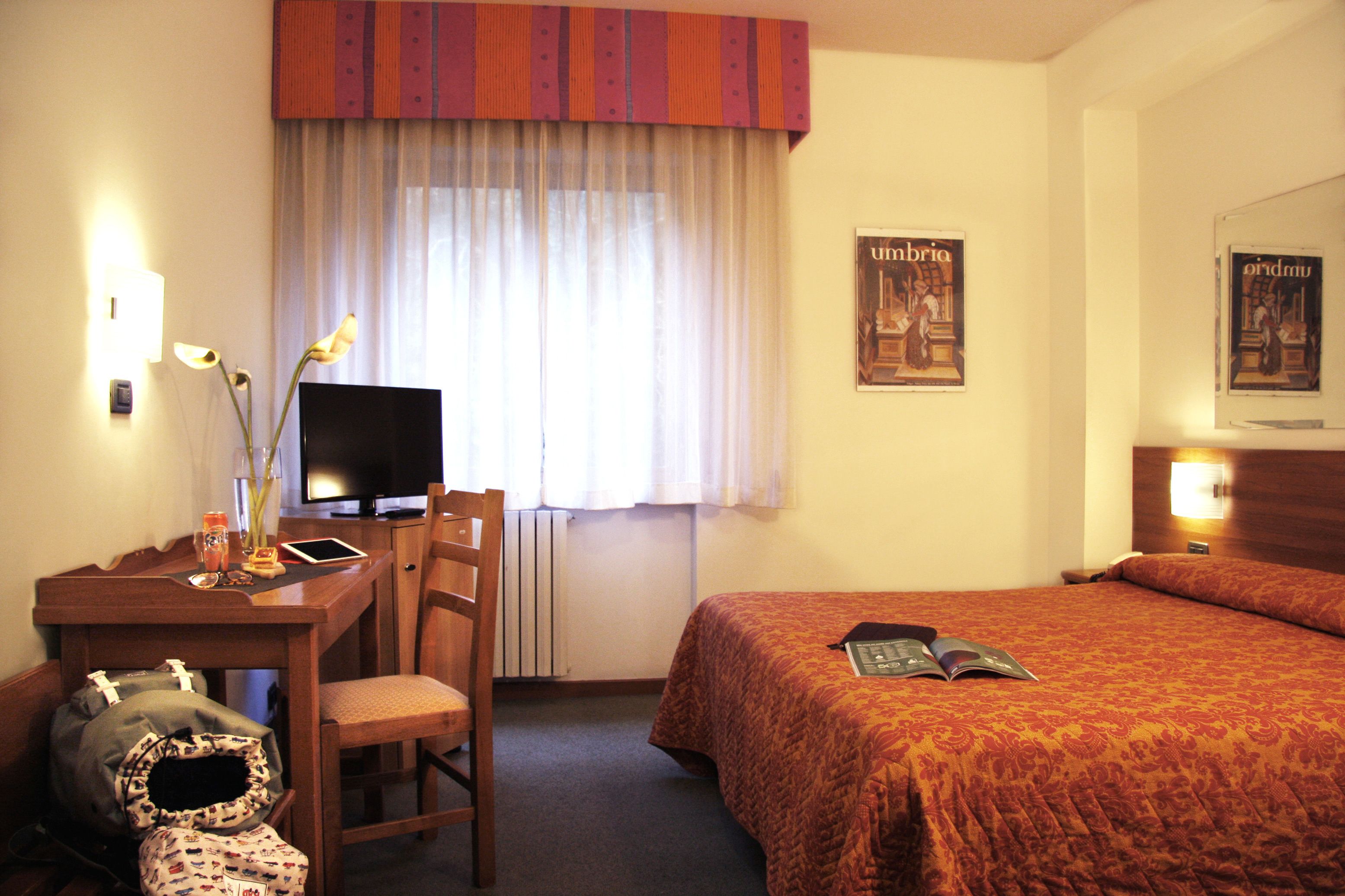 A hotel room with a bed, a desk with a computer, a chair, and a radiator.
