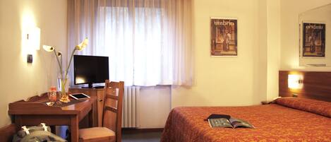 A hotel room with a bed, a desk with a computer, a chair, and a radiator.