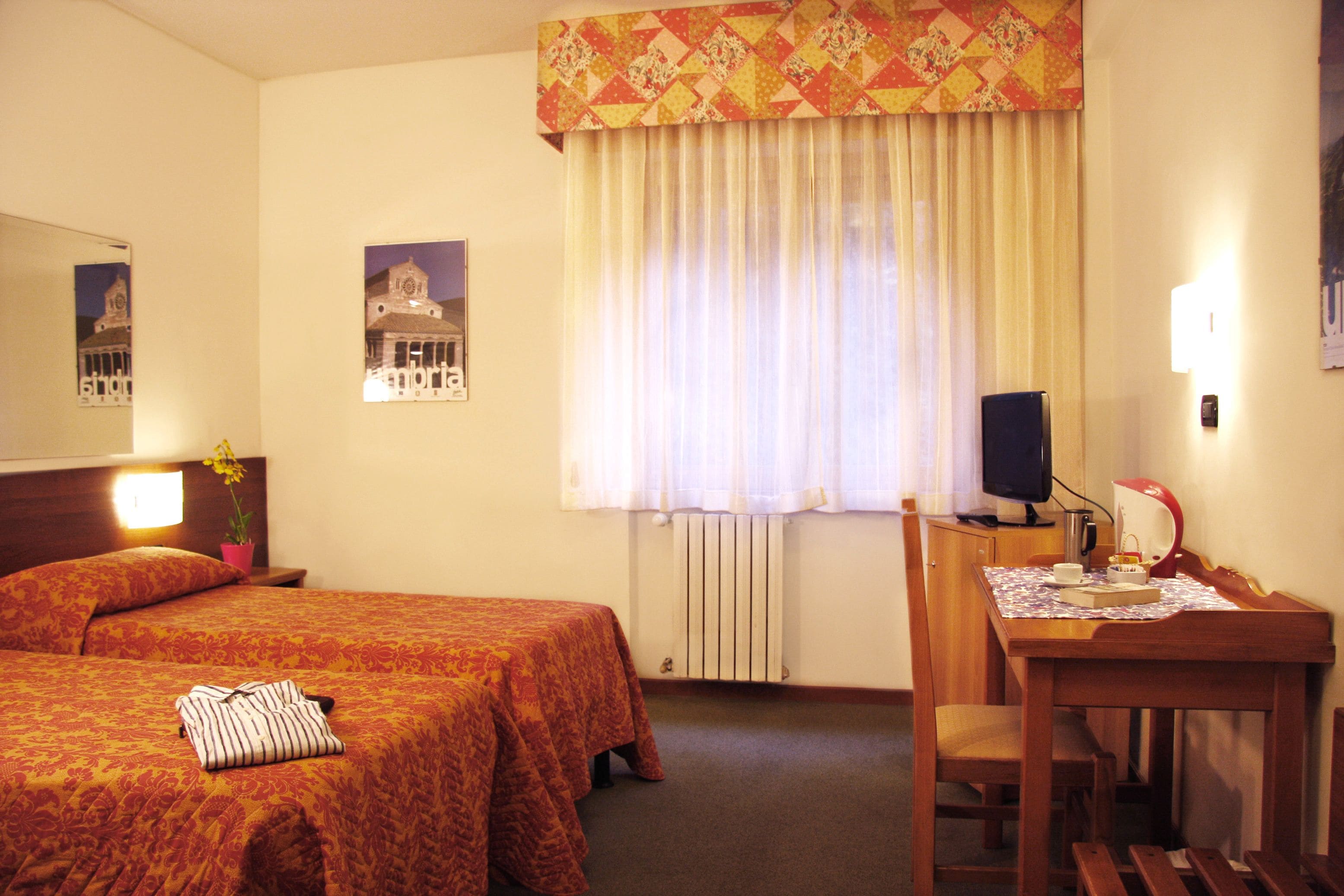 Habitación de hotel con dos camas, un escritorio con silla, un radiador y una ventana con cortinas.