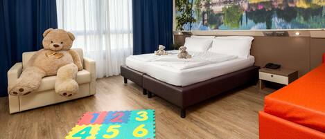 A hotel room with a bed, a chair with a teddy bear, a colorful number mat on the floor, and a scenic wall mural.