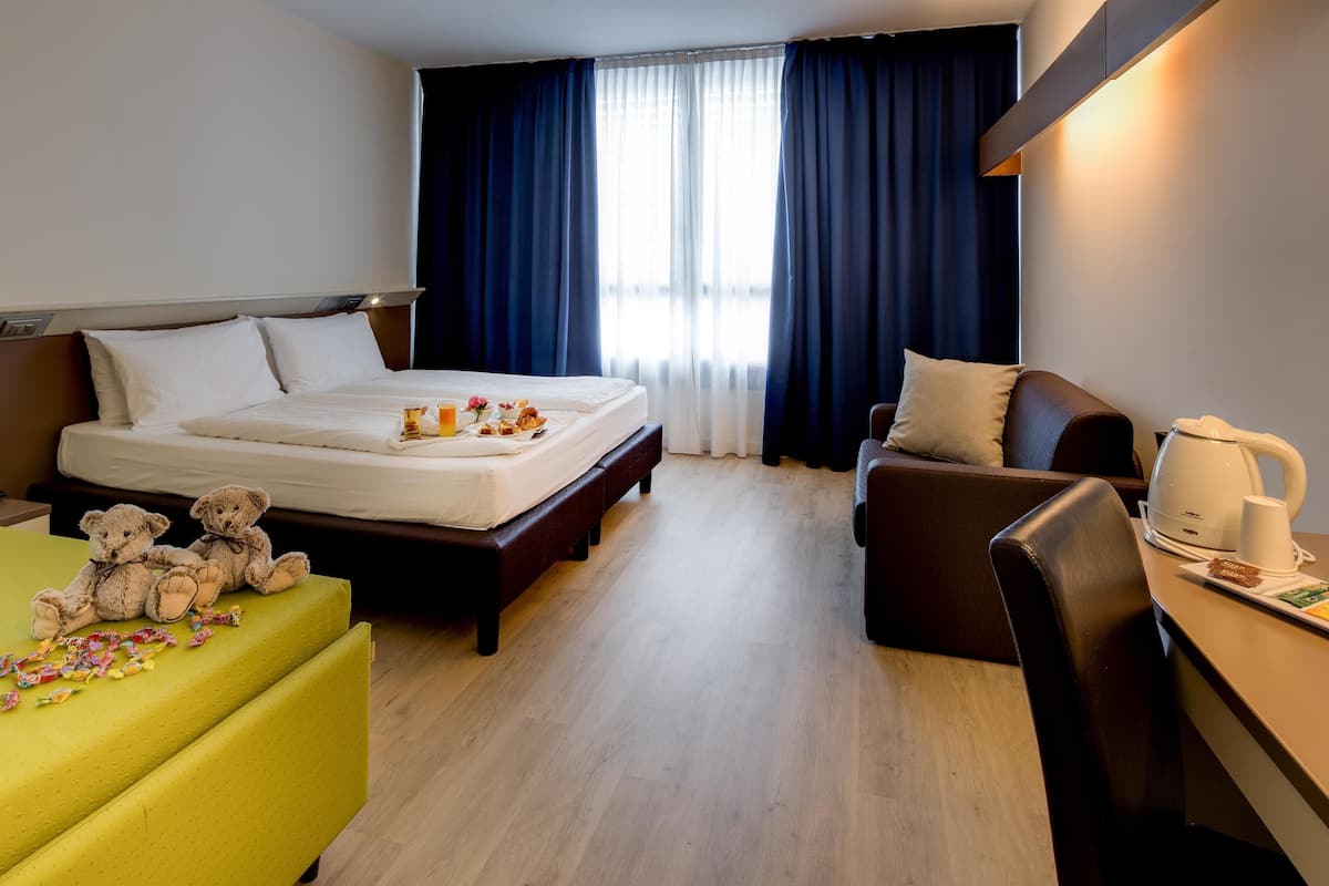 A hotel room with a bed, a sofa, a desk with a kettle, and a teddy bear on the sofa.