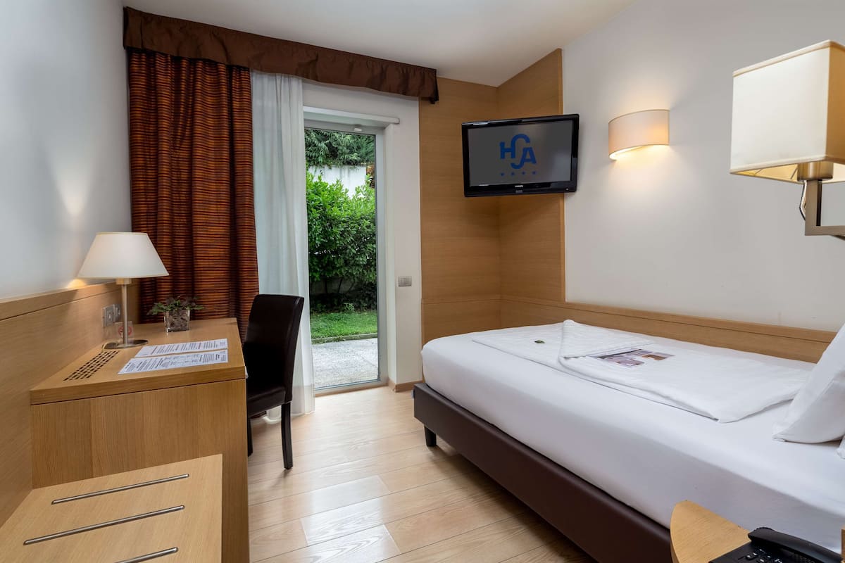 A hotel room with a bed, a desk with a lamp, a TV mounted on the wall, and a window with curtains.