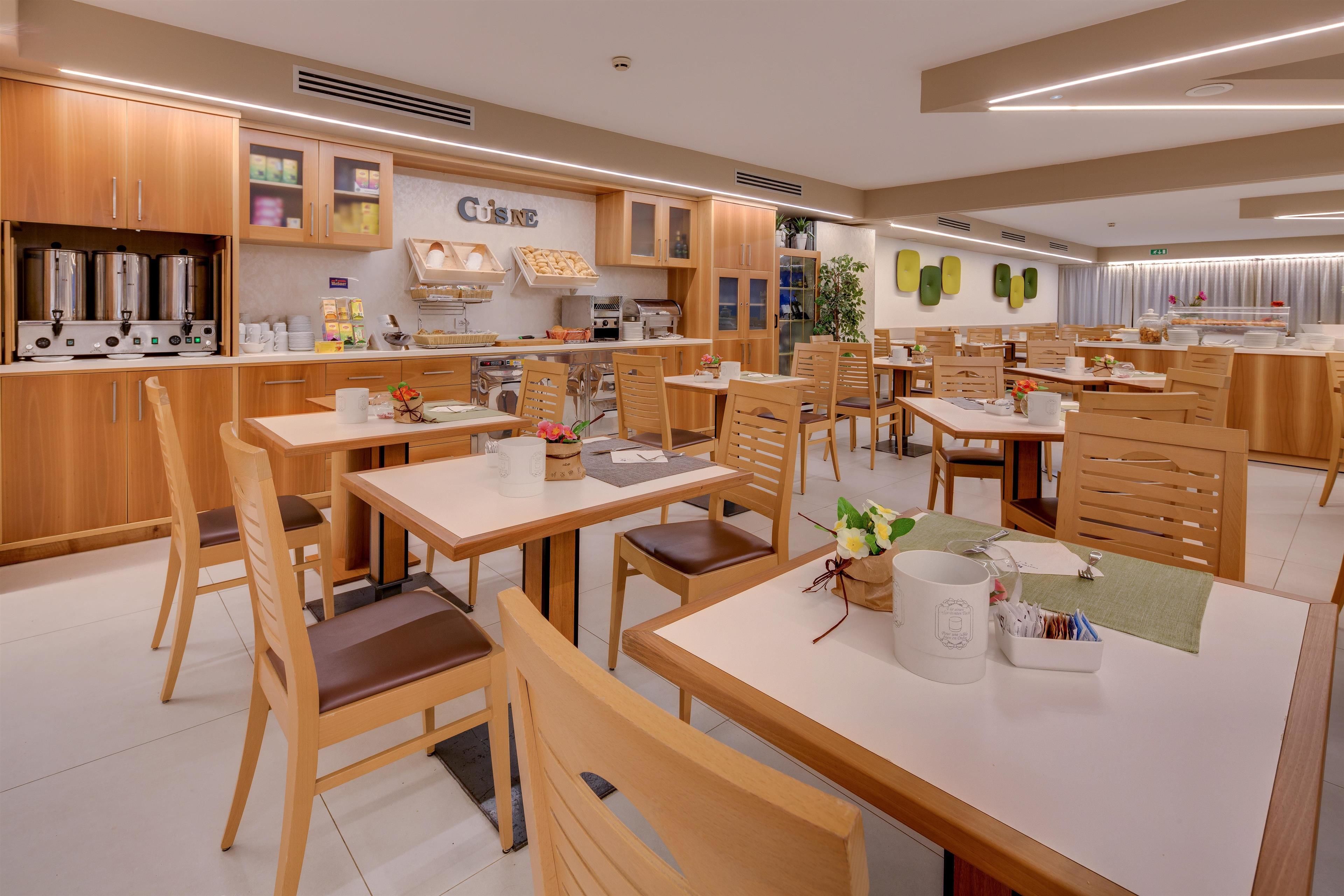 Un espace repas moderne avec des tables et des chaises en bois, un coin cuisine équipé d’appareils électroménagers en acier inoxydable, et un mur orné d’une enseigne « CUSNC ».